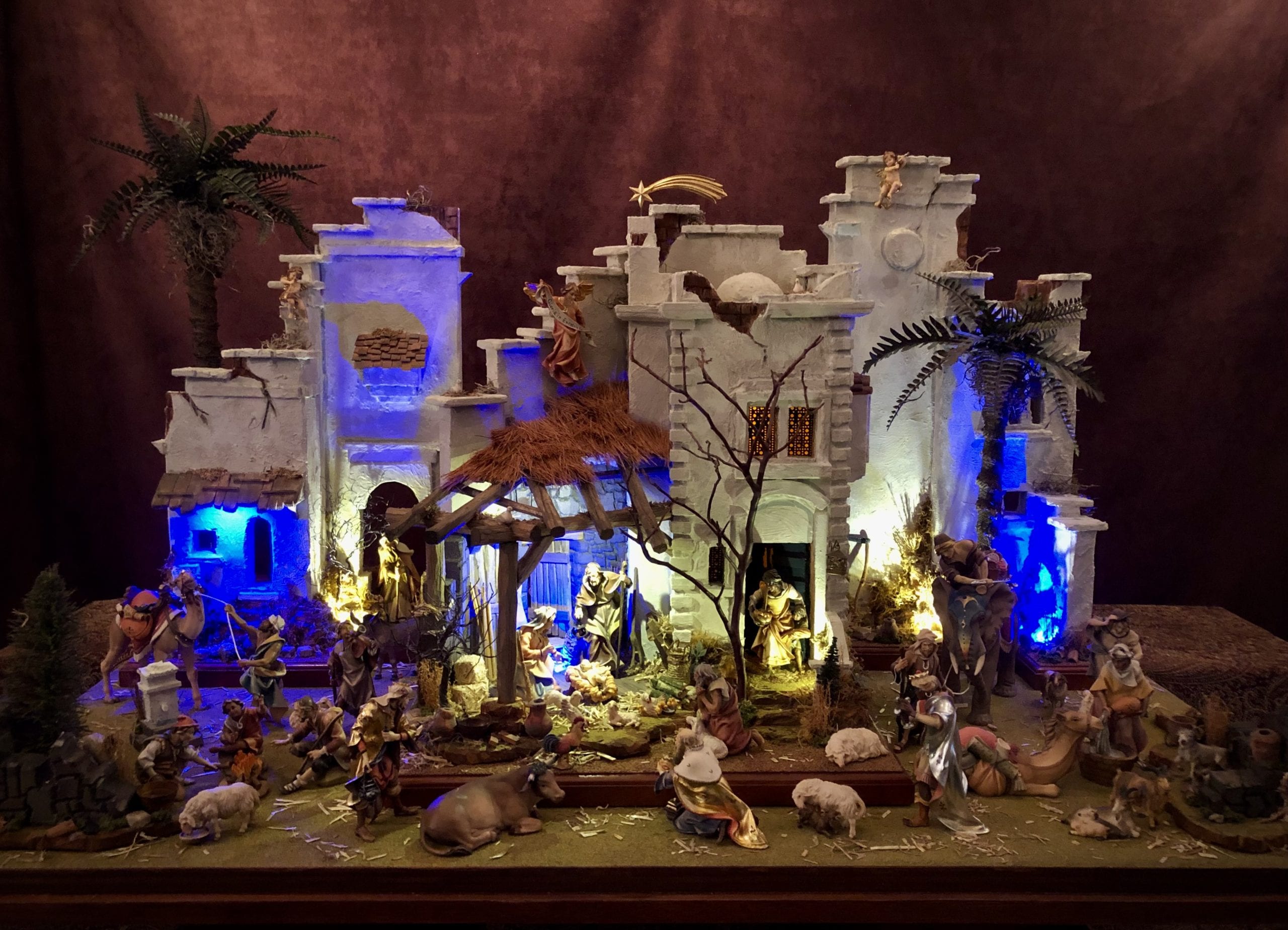 One-of-a-kind illuminated nativity village of whitewashed buildings and palm trees, glowing blue and gold against a dark backdrop.