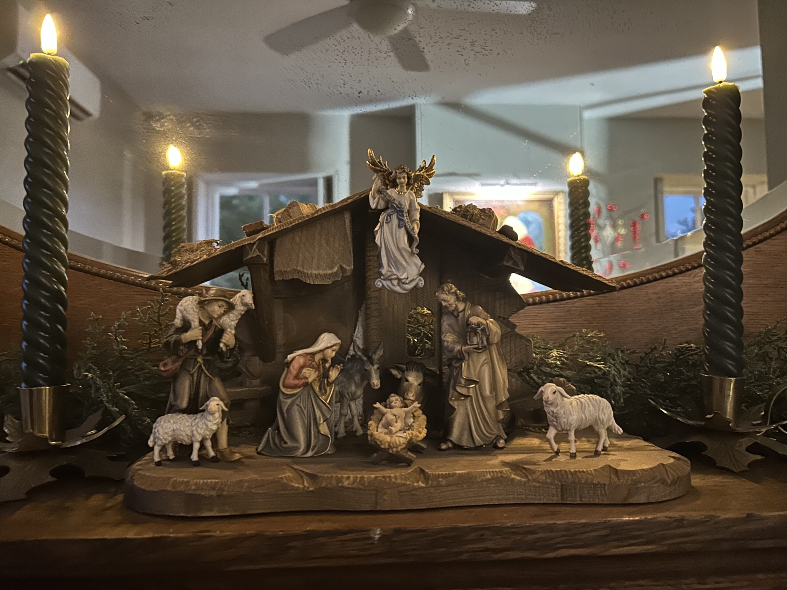 Carved wooden stable with the Holy Family on a mantel, flanked by evergreen boughs and tall candles.
