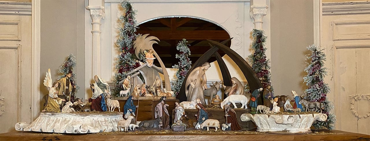 Antique nativity figures arranged across a mantel between frosted Christmas trees in a plasterwork interior.