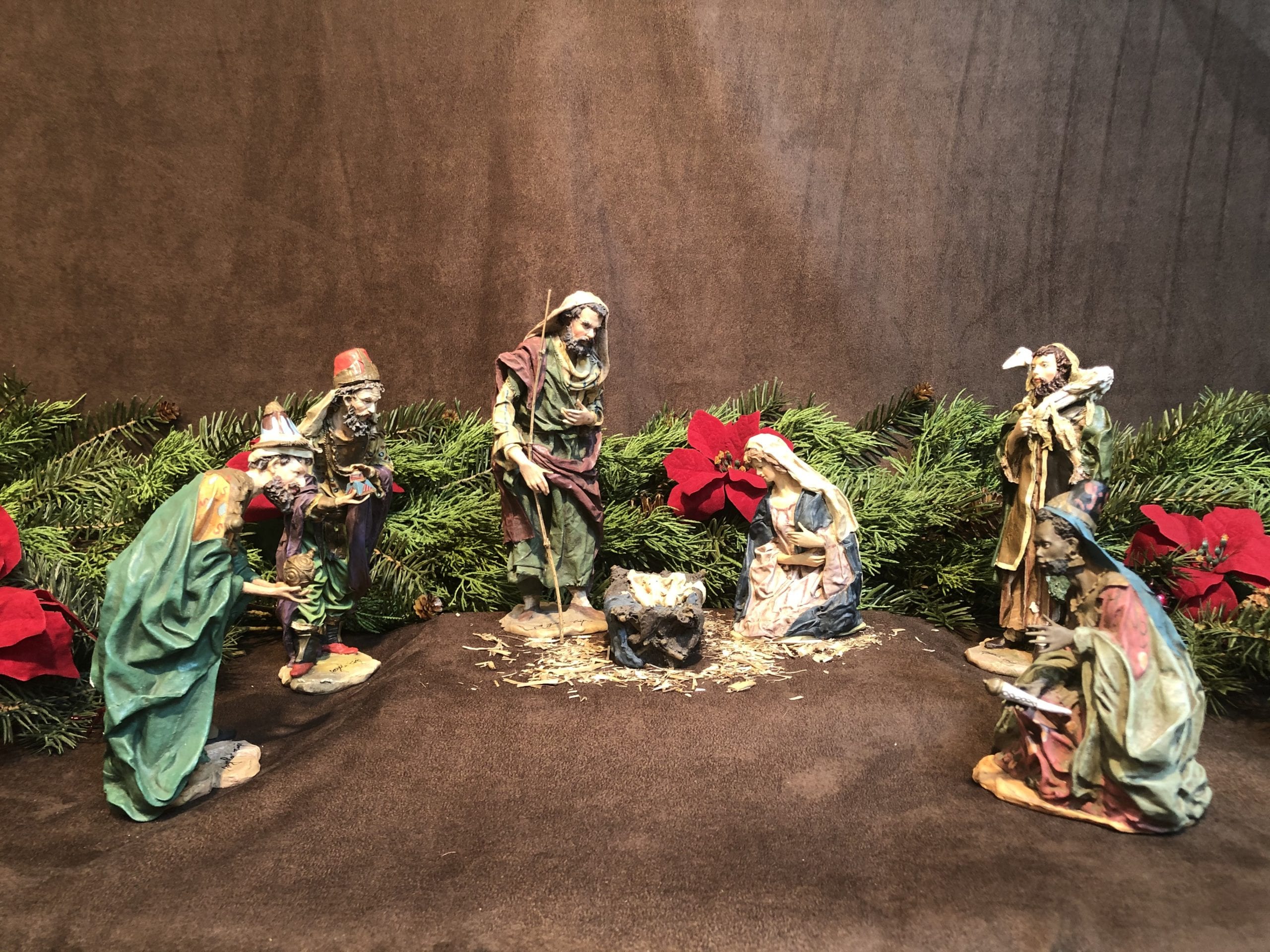Full Angela Tripi Nativity set with Mary, Joseph, Baby Jesus in manger, shepherds, and kings on brown background with greenery and red poinsettias